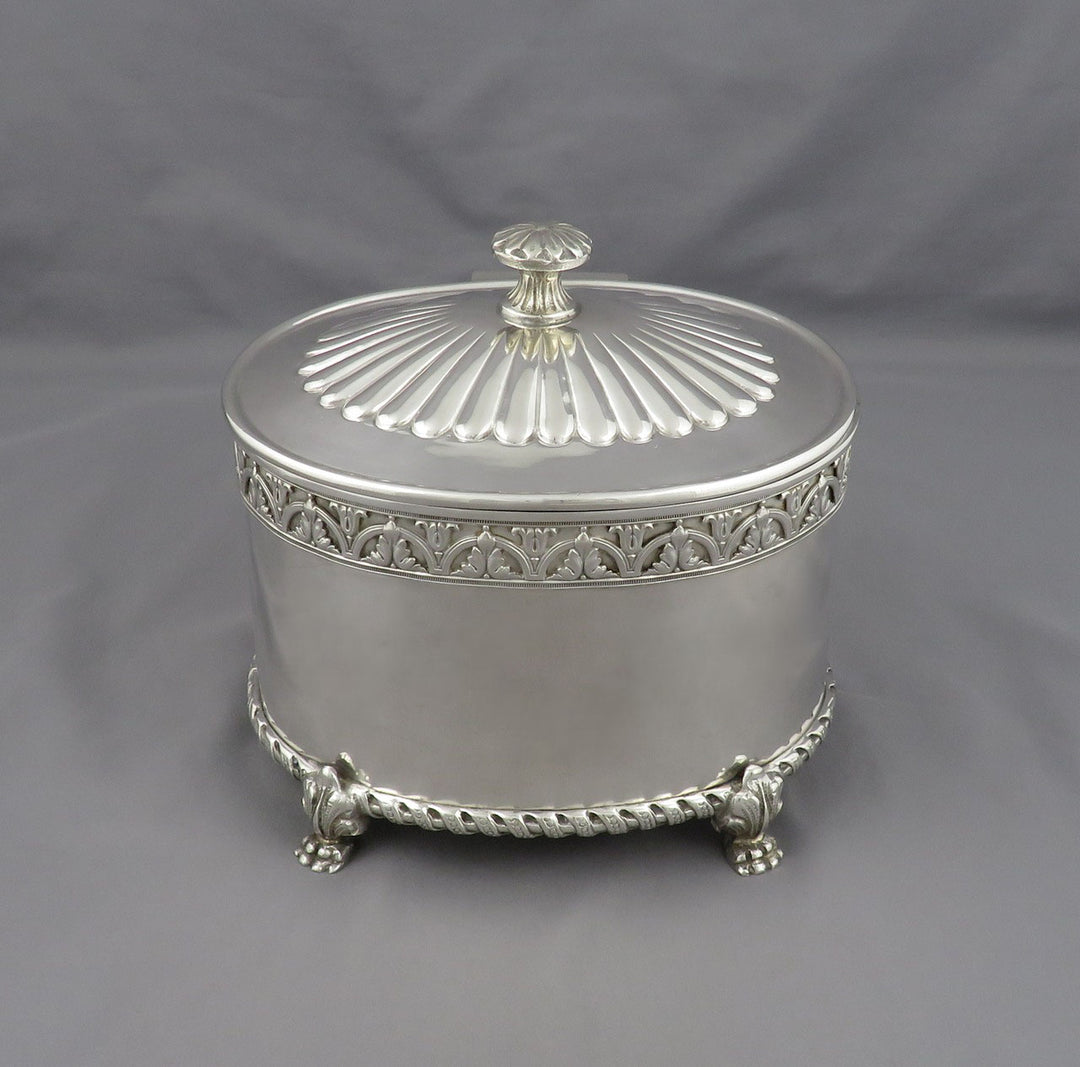 An ornate silver biscuit jar or caddy with decorative trim, a ribbed lid, and small feet, photographed against a plain gray fabric backdrop.