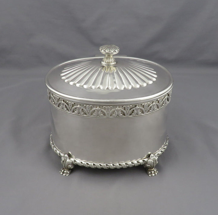 An ornate silver biscuit jar or caddy with decorative trim, a ribbed lid, and small feet, photographed against a plain gray fabric backdrop.