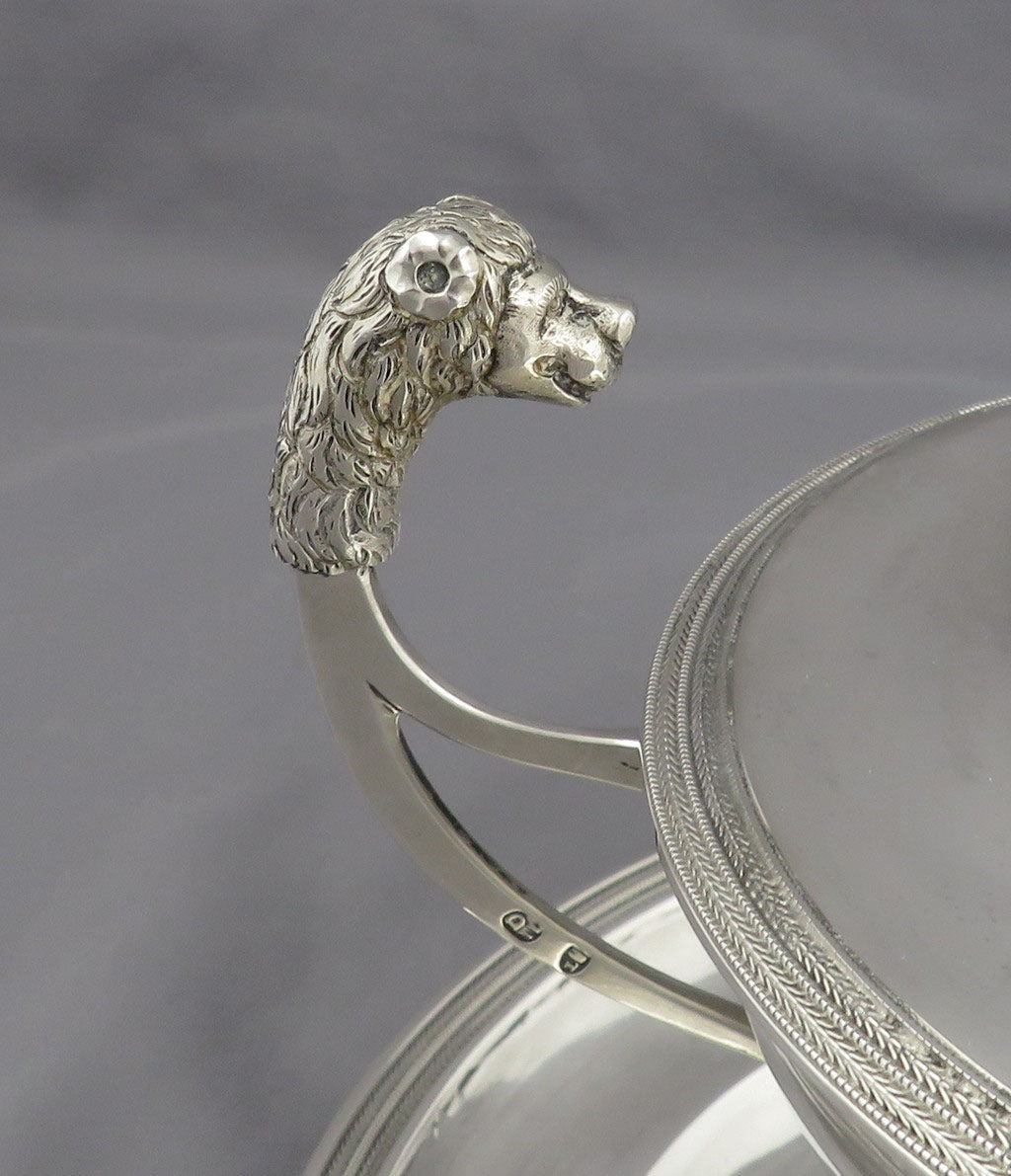 Close-up of a silver sauce tureen with a sculpted lion head handle and a decorative engraved rim on a matching round base, photographed against a soft gray background.