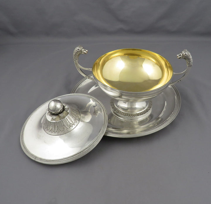 An antique silver sauce tureen with a polished gold interior, featuring decorative lion handles and a matching silver lid resting beside it on a gray fabric background.