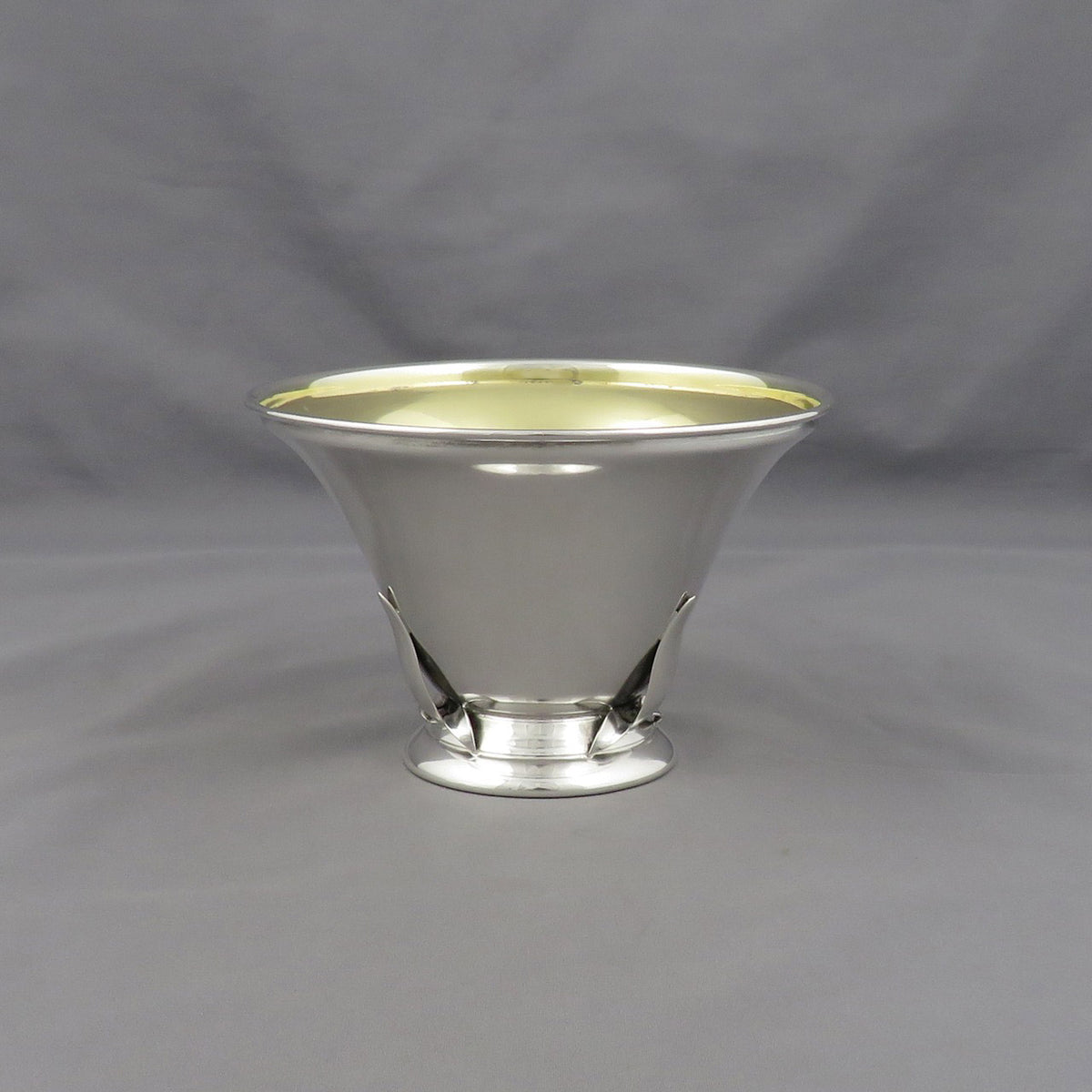 A polished silver pedestal bowl with a flared rim and a small base, photographed against a neutral gray fabric backdrop.