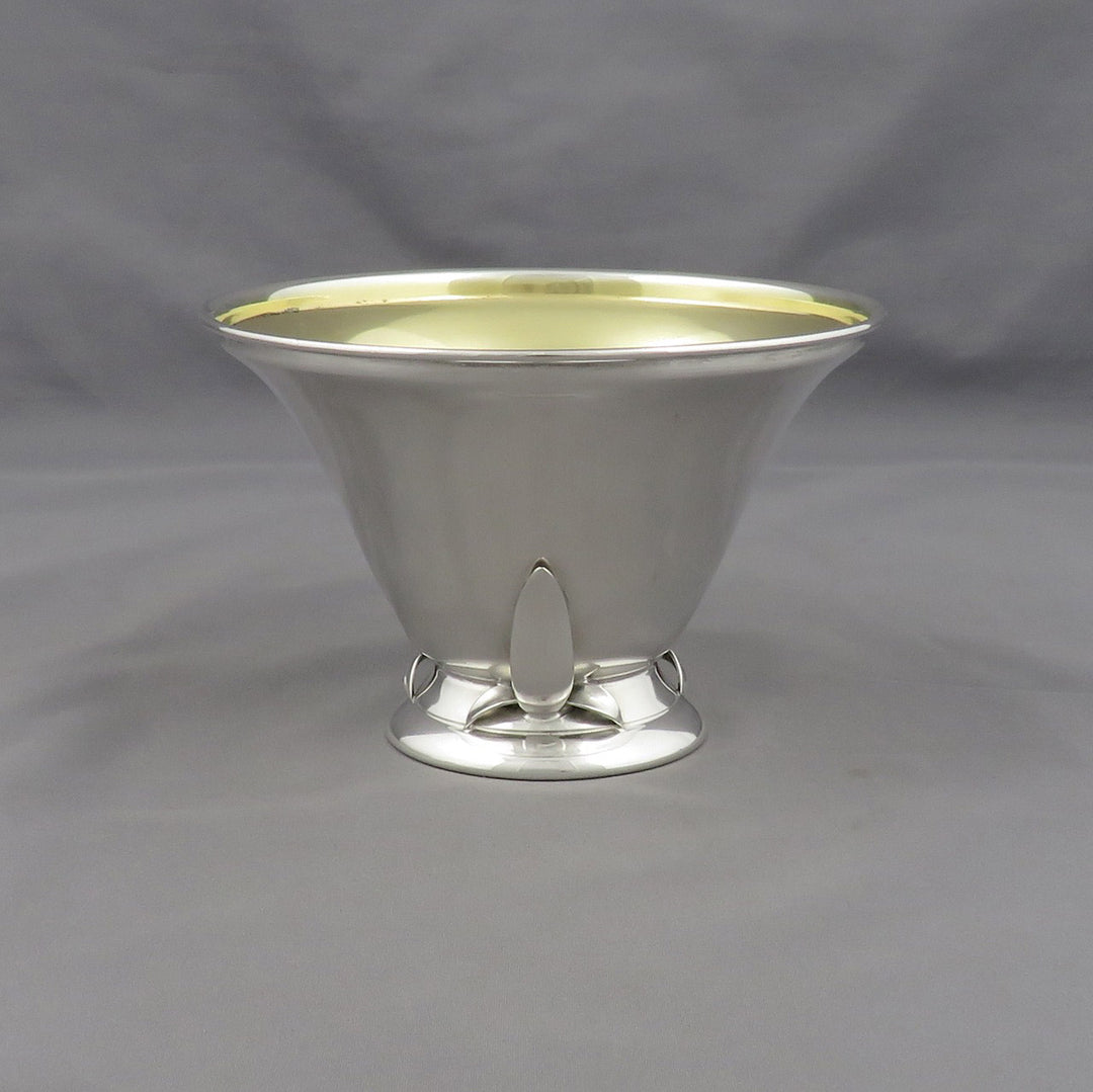 A polished silver pedestal bowl with a flared rim, a small central pedestal foot, and a pale gold interior, photographed against a neutral gray fabric backdrop.