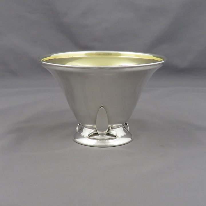 A polished silver pedestal bowl with a flared rim, a small central pedestal foot, and a pale gold interior, photographed against a neutral gray fabric backdrop.