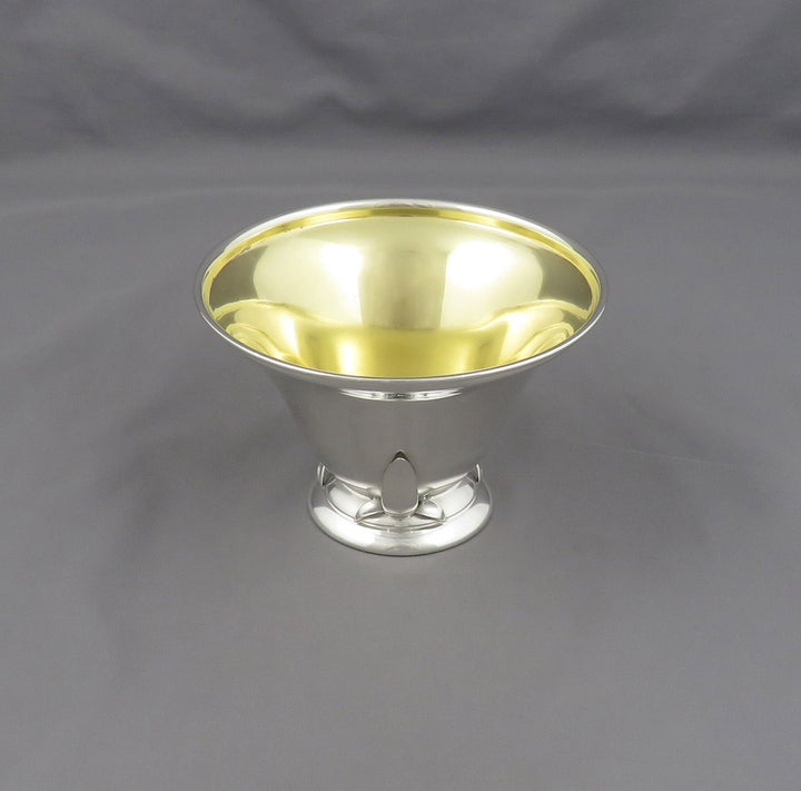 A polished silver pedestal bowl with a highly reflective gold-toned interior, set against a neutral gray fabric backdrop.