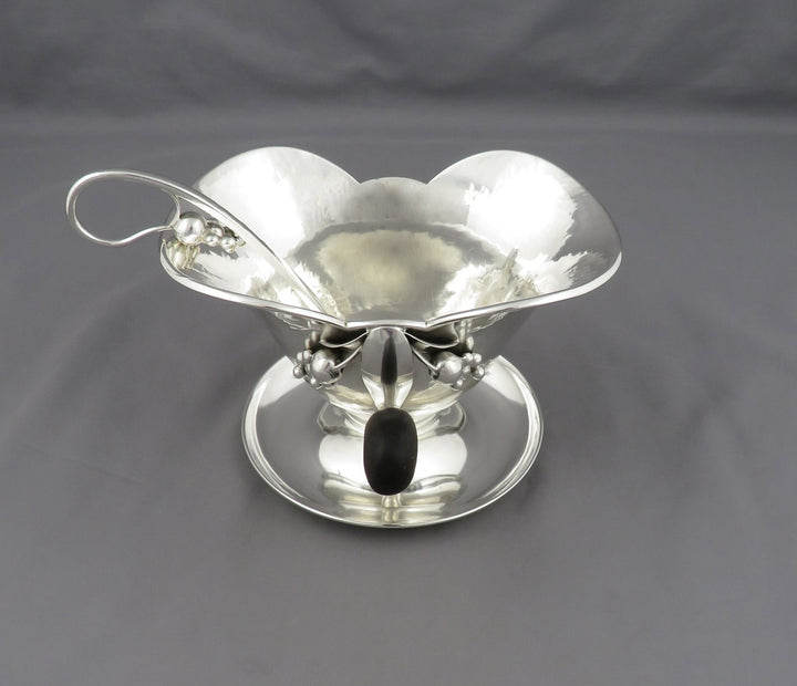 A polished silver gravy boat by Poul Petersen with a scalloped rim, a side handle, and a matching saucer, photographed on a dark gray fabric background.