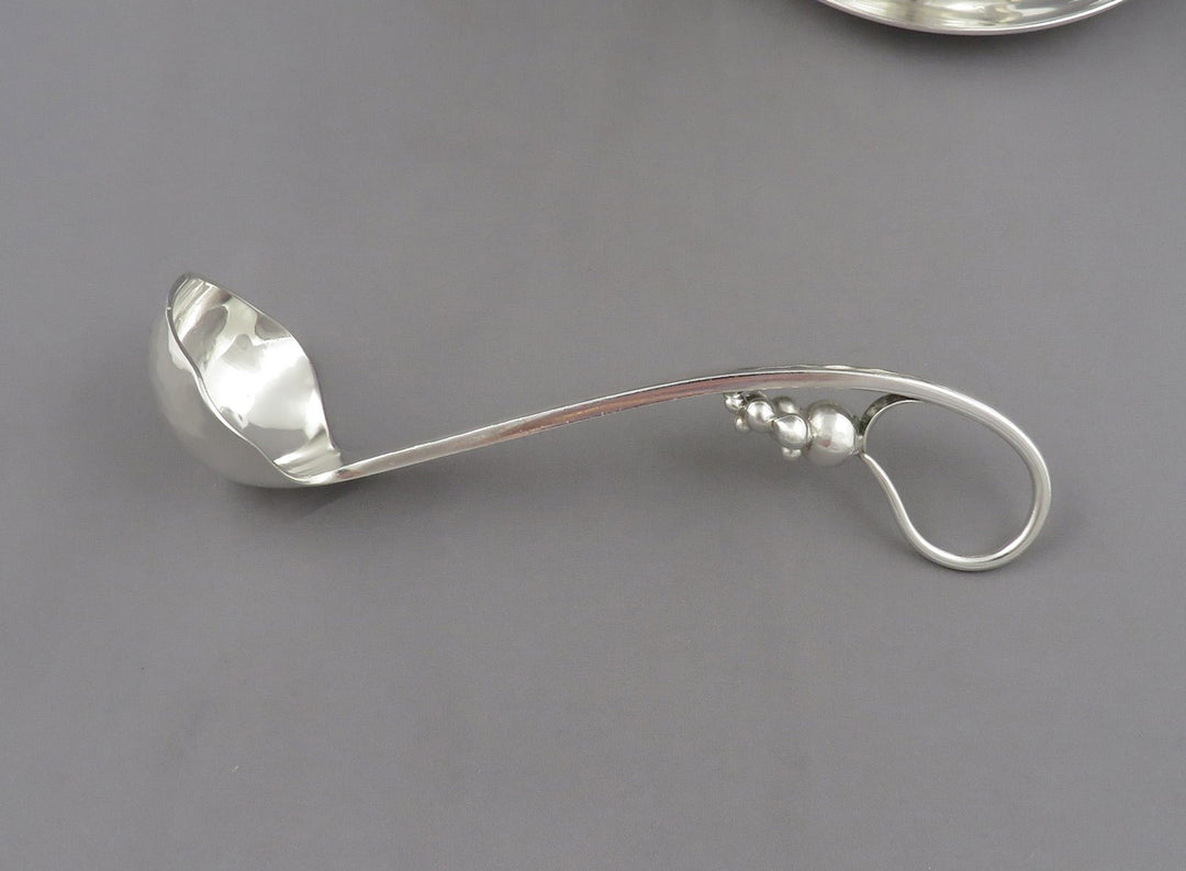A polished silver ladle by Poul Petersen with a decorative loop handle, photographed on a smooth gray surface.