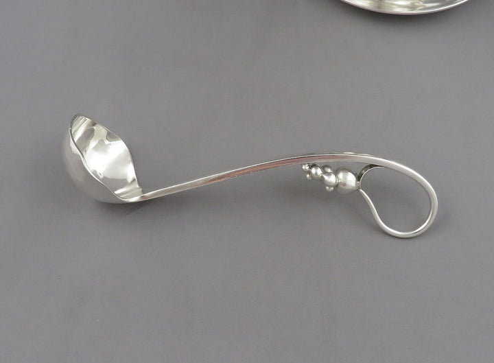 A polished silver ladle by Poul Petersen with a decorative loop handle, photographed on a smooth gray surface.