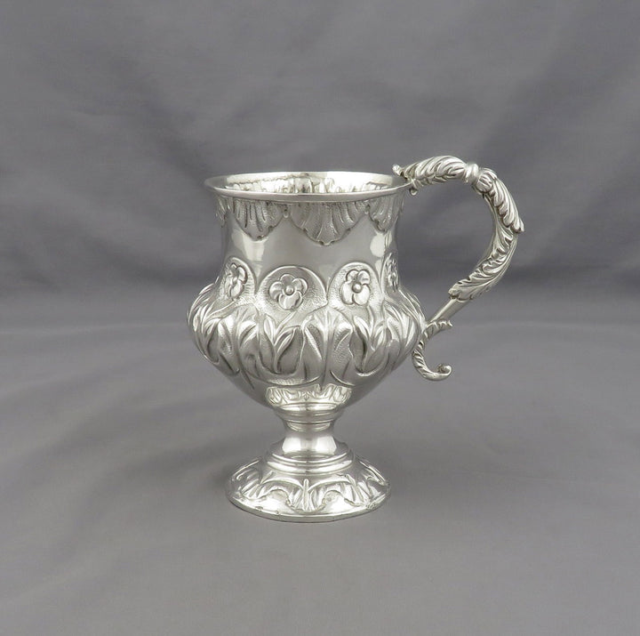 An ornate sterling silver footed cup with a decorative handle, featuring embossed floral and scrollwork patterns, set against a neutral gray background.